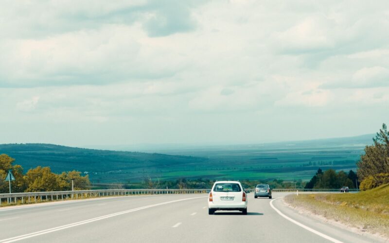A serene view of a road with cars, bordered by lush landscapes under a cloudy sky.