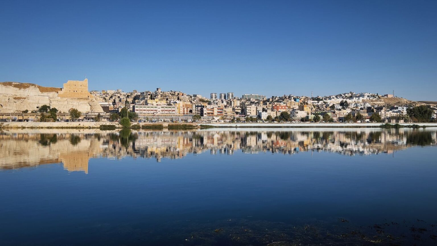Panoramic view of a vibrant cityscape reflecting on a tranquil body of water under a clear blue sky.