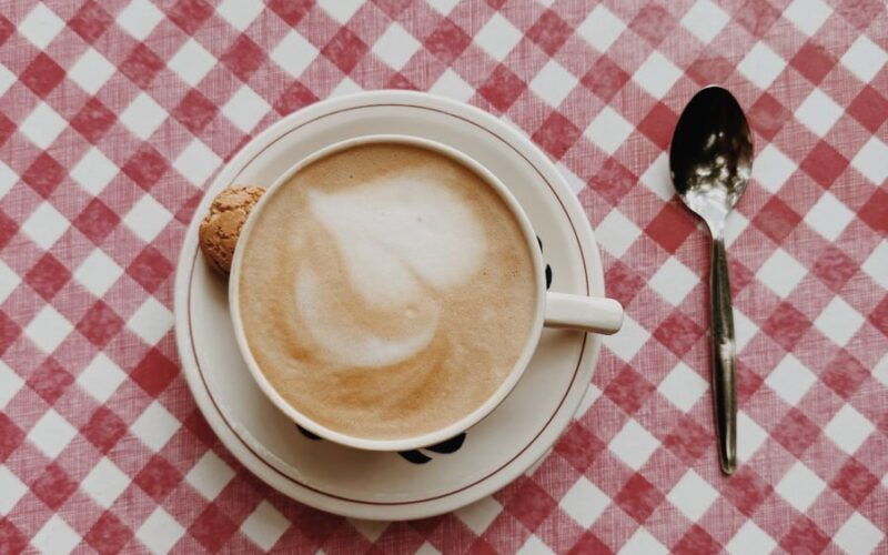 A cappuccino cup with a cookie on a red checkered tablecloth, with a spoon beside it.